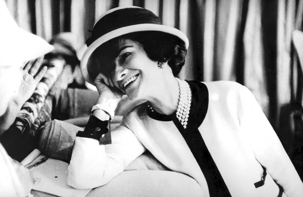 Gabrielle Chanel dressed in a tweed jacket and hat and smiling leaning on her hand